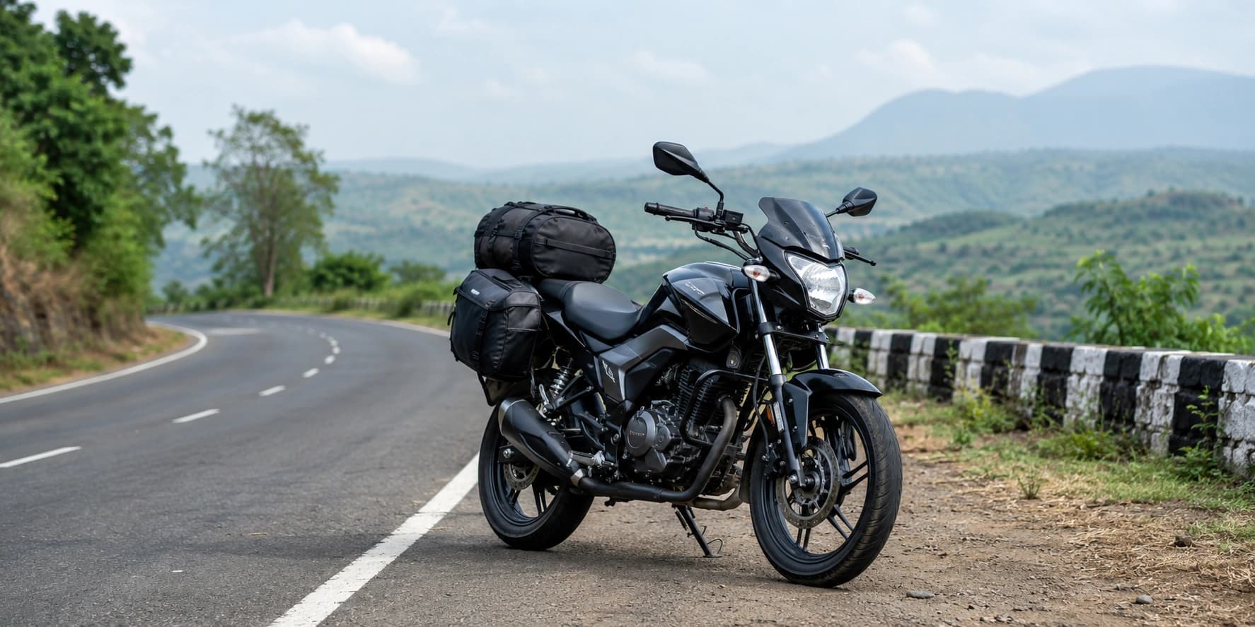 A modern black touring motorcycle with luggage parked beside a winding scenic road through green hills in India.